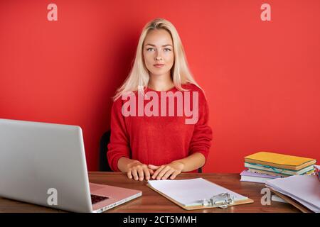 Porträt der jungen kaukasischen blonde Frau am Arbeitsplatz, sitzt sie in lässiger Kleidung am Schreibtisch, genießen Sie die Arbeit auf Laptop und mit Dokumenten. Isoliert Stockfoto