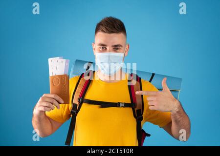 Reisen während einer Pandemie. Junger Mann mit Rucksack trägt Maske, zeigt auf Pass und Flugtickets blauen Hintergrund Stockfoto