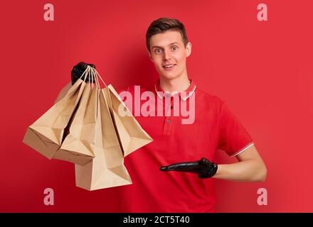 Junge kaukasische Delivery Mann Mitarbeiter in roten T-Shirt Uniform und in Schutzhandschuhen halten Handwerk Papierpaket mit Lebensmitteln isoliert auf rotem Hintergrund, Stockfoto