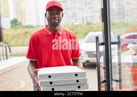 Portrait von aufgeschlossenen afican deliveryman mit Pizza in den Händen, gehen, um es für Kunden, Blick auf Kamera tragen rote Uniform Stockfoto