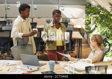 Porträt von zwei zeitgenössischen afroamerikanischen Menschen in lässiger Kleidung im Gespräch mit Kollegen während der Arbeit im Büro, Kopierer Raum Stockfoto