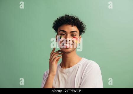 Mann mit Regenbogen Gesichtsfarbe feiert Homosexuell Stolz. Gay Mann auf grauem Hintergrund stehend Blick auf Kamera und lächelnd. Stockfoto