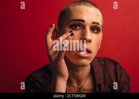 Transgender männlich legte Make-up auf vor rotem Hintergrund. Mann mit Make-up und Nase Piercing Blick auf Kamera. Stockfoto