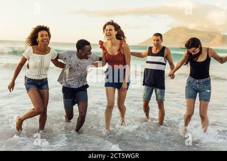 Freunde im Urlaub spielen im Meer. Multiethnische Männer und Frauen Freunde laufen im Meerwasser halten sich gegenseitig. Stockfoto