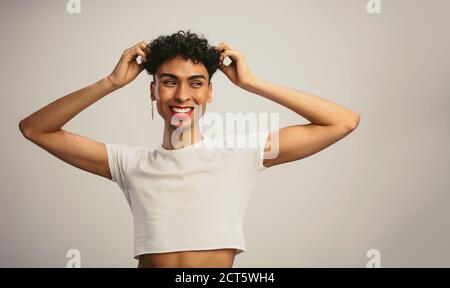 Geschlecht Flüssigkeit Mann trägt Crop Top mit seinen Händen im Haar vor weißem Hintergrund. Gay Mann schaut weg und lächelt. Stockfoto