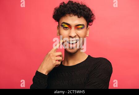 Gay Mann trägt bunte Schatten auf den Augenlidern zwinkert bei der Kamera. Glücklich Transgender männlich winken ein Auge vor rotem Hintergrund. Stockfoto