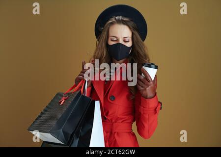 Leben während covid-19 Pandemie. Entspannte 40 Jahre alte Frau in rotem Mantel mit schwarzer Maske, Tasse und Papier Einkaufstaschen vor Bronze Hintergrund. Stockfoto