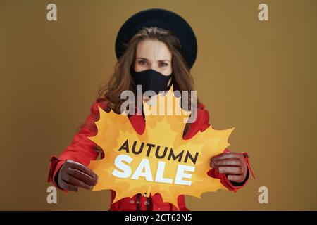 Hallo Herbst. Nahaufnahme einer Frau mittleren Alters im roten Mantel mit Herbstverkauf Blatt Form Banner und schwarze Maske auf braunem Hintergrund. Stockfoto