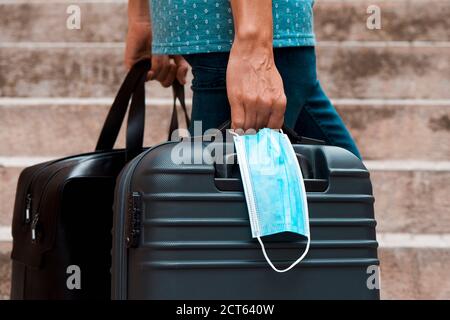 Nahaufnahme eines jungen Mannes auf der Straße, in legerer Kleidung, mit einer schwarzen Reisetasche in der Hand und einem grauen Koffer und einer Einweg-Gesichtsmaske in der ot Stockfoto