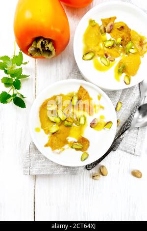 Dessert aus Joghurt, Kaki und Honig mit Vanille, Kardamom und Pistazien in zwei Schüsseln auf Serviette, Minze und Löffel auf dem Hintergrund von hellem Holz Stockfoto