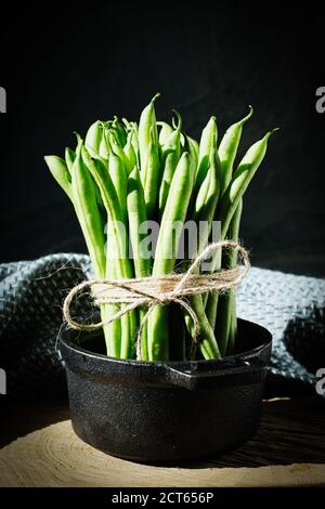 Frisch geerntete Bohnen, gebunden mit einer Schnur in einem gusseisernen Topf auf einem Holzteller mit schwarzem Hintergrund Stockfoto