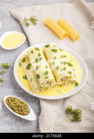 Cannelloni-Pasta mit Eiersauce, Frischkäse und Oregano-Blättern auf grauem Betonboden mit Leinentextilien. Seitenansicht, Nahaufnahme. Stockfoto