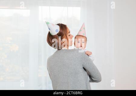 Eine junge Mutter hält ein niedliches Baby in den Armen. Festliche Papierhüte auf ihren Köpfen. Rückansicht. Fenster im Hintergrund. Konzept der Mutterschaft und glücklich Stockfoto