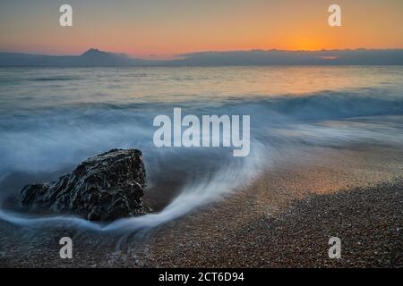 Ein Felsen steht auf dem Kiesstrand und wird von der Meereswelle bei Sonnenuntergang mit sehr schönen Farben des Meereshimmels der Welle und der Kieselsteine getroffen. Die Welle Stockfoto