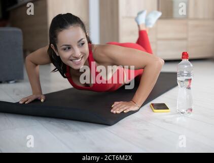 Junge Frau im roten Trainingsanzug macht zu Hause Sport oder Yoga. Fröhliche positive und glückliche Mädchen stehen auf Plank Position auf den Knien. Lehnen Sie sich auf die Hände und Stockfoto