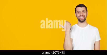 Junger Mann mit Bart in weißem T-Shirt glückliches Gesicht lächelnd mit glücklichem Gesicht suchen und zeigen auf die Seite mit Daumen nach oben. Steht auf isoliertem gelben Hintergrund. Stockfoto