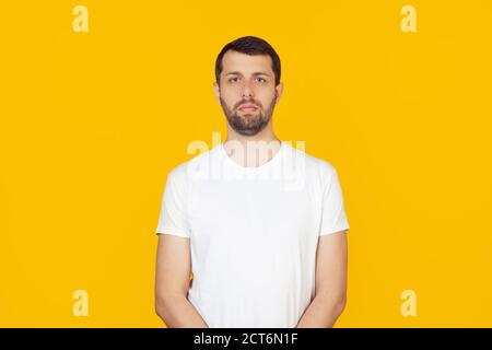 Junger Mann mit einem Bart in einem weißen T-Shirt mit einem ernsthaften Gesichtsausdruck. Einfacher und natürlicher Blick in die Kamera. Steht auf isoliertem gelben Hintergrund. Stockfoto