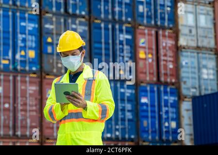 Container-Arbeiter tragen Schutzmaske gegen Covid-19 arbeiten und halten digitale Tablette auf logistischen Versand Fracht Container Hof zu schützen. Stockfoto