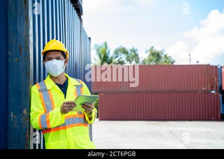Container-Arbeiter tragen Schutzmaske gegen Covid-19 arbeiten und halten digitale Tablette auf logistischen Versand Fracht Container Hof zu schützen. Stockfoto