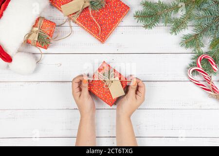 Weibliche Hände öffnen eine Geschenkbox Verpackung in das rote Papier, flach legen. Vorbereitung auf die Weihnachtsferien. Draufsicht der Hände auf Holztisch mit Tannenbaum Stockfoto