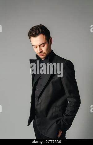 Portrait einer jungen gutaussehenden Mann in einem schwarzen Anzug, stehen und nach unten schaut, Hand in der Tasche, gegen normales studio Hintergrund Stockfoto