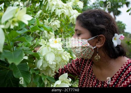 Schöne Dame mit Blumenmaske, Ohrringe und Designer-Top duftet weißen Mussaenda Blumen. Schutz vor Corona auf stilvolle Weise. Stockfoto