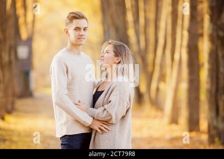 Liebevolles Paar steht umarmt im Herbstwald in hellbeigen Pullovern, gelber Hintergrund mit Blättern Stockfoto