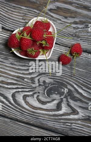 Himbeeren mit Schwänzen liegen in einer herzförmigen Untertasse. Mehrere Beeren sind in der Nähe. Auf schwarzen Brettern, mit einer ausdrucksstarken holzigen Textur. Stockfoto