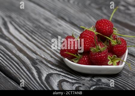 Himbeeren mit Schwänzen liegen in einer herzförmigen Untertasse. Auf schwarzen Brettern, mit einer ausdrucksstarken holzigen Textur. Stockfoto