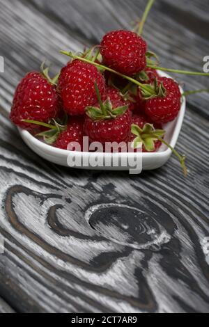 Himbeeren mit Schwänzen liegen in einer herzförmigen Untertasse. Auf schwarzen Brettern, mit einer ausdrucksstarken holzigen Textur. Stockfoto