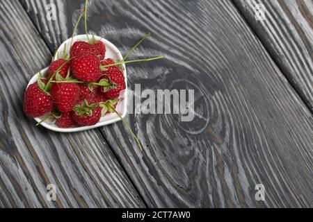 Himbeeren mit Schwänzen liegen in einer herzförmigen Untertasse. Auf schwarzen Brettern, mit einer ausdrucksstarken holzigen Textur. Stockfoto