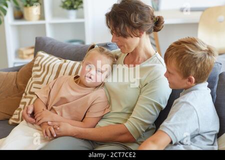 Portrait der modernen Mutter umarmen Mädchen mit Down-Syndrom, während Sitzen auf der Couch mit zwei Kindern in der häuslichen Einrichtung Stockfoto