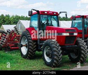 Leuchtend rote Tasche Traktor mit Mähwerkzeug auf der Rückseite geparkt. Im Hintergrund befinden sich Gewächshäuser, Bäume und ein strahlend blauer Himmel. Stockfoto