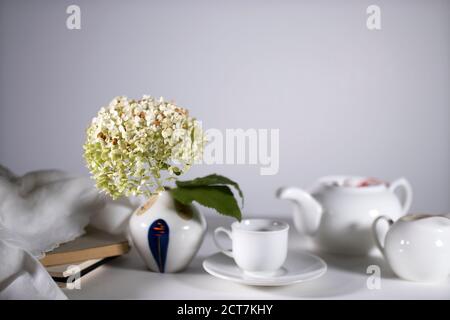 Weiße Teekanne, Tasse und Untertasse und Milchkännchen zum Teetrinken. Grüne getrocknete Hortensien in einer kleinen Steingutvase im Stil der siebziger Jahre schmückt die Stockfoto