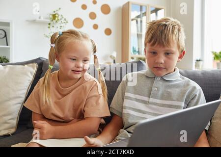 Portrait von niedlichen Mädchen mit Down-Syndrom lächelnd beim Blick Am Laptop-Bildschirm und sitzen auf dem Sofa mit Bruder an Zu Hause Stockfoto