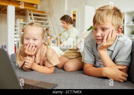 Portrait von niedlichen Mädchen mit Down-Syndrom lächeln glücklich, während Blick auf Laptop-Bildschirm und liegend auf dem Sofa mit Bruder Zu Hause Stockfoto
