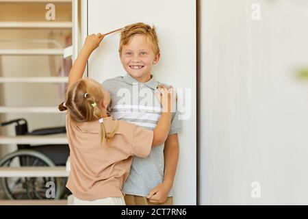 Portrait von niedlichen Mädchen Messung Höhe der glücklichen älteren Bruder steht gegen weiße Wand zu Hause, kopieren Raum Stockfoto