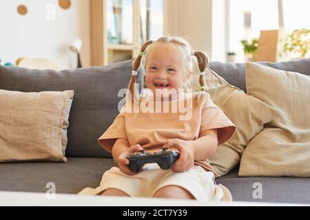 Portrait von niedlichen Mädchen mit Down-Syndrom spielen Videospiele und glücklich lachen, während auf der Couch im Wohnzimmer sitzen, kopieren Raum Stockfoto