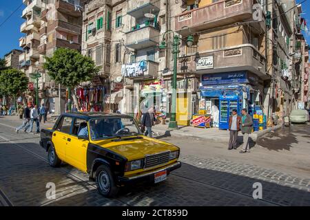 Ein Taxi fährt eine Straße entlang an Wohngebäuden in einem Vorort von Alexandria in Ägypten vorbei. Stockfoto