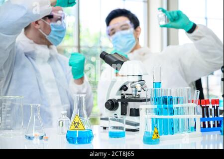 Laborgeräte mit blauer Flüssigkeit im Inneren stehen auf dem Tisch Mit paar männlichen Wissenschaftler tragen Schutzanzug hält Impfstoff für Forschung im Labor Stockfoto