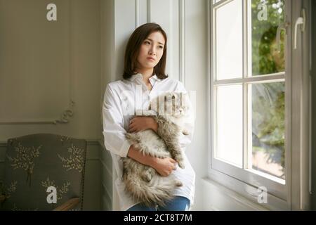 Schöne junge asiatische Frau stehen am Fenster zu Hause halten Eine Katze in den Armen, die deprimiert und traurig aussieht Stockfoto