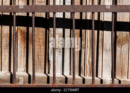 Ein alter, rostig geschmiedeter Zaun. Die Textur eines massiven Metallzauns. Hinter dem Zaun befindet sich ein Holzhintergrund, auf den der Schatten fällt. Stockfoto