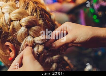 Prozess der Flechtarbeiten der Meister webt Zöpfe auf ihrem Kopf blonde kleine Mädchen in Schönheitssalon Nahaufnahme. Professionelle Haarpflege und Frisuren erstellen. Stockfoto