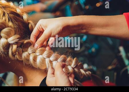 Prozess der Flechtarbeiten der Meister webt Zöpfe auf ihrem Kopf blonde kleine Mädchen in Schönheitssalon Nahaufnahme. Professionelle Haarpflege und Frisuren erstellen. Stockfoto