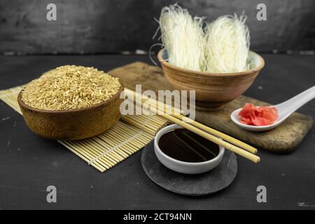 Brauner Reis in einer Holzschüssel, asiatischer Küchenhintergrund mit Bambusmatte, Essstäbchen, Sojasauce und Glasreis-Nudeln auf schwarzem Hintergrund Stockfoto