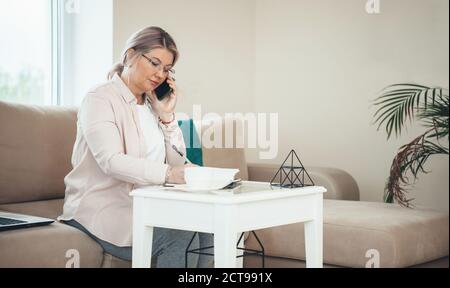 Seitenansicht Foto einer Geschäftsfrau, die am Telefon spricht Während Sie etwas schreiben und auf der Couch in der Nähe des sitzen Notebook Stockfoto