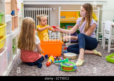 Junge weibliche Therapeutin trägt schützende Gesichtsmaske spielt mit zwei Kleinkindern Mädchen während der Ergotherapie Kind. Stockfoto