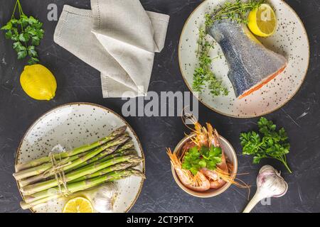 Forellen umgeben Petersilie, Zitrone, Garnelen, Garnelen, Spargel in Keramikplatten. Tischoberfläche aus schwarzem Beton. Gesunde Meeresfrüchte Hintergrund. Stockfoto