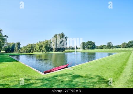 Ein Wasserbrunnen und ein überdachtes Boot im großen Teich (Grand Etang) auf dem Gelände von Duke of Buccleuch's Boughton House, Kettering, Northamptonshire. Stockfoto