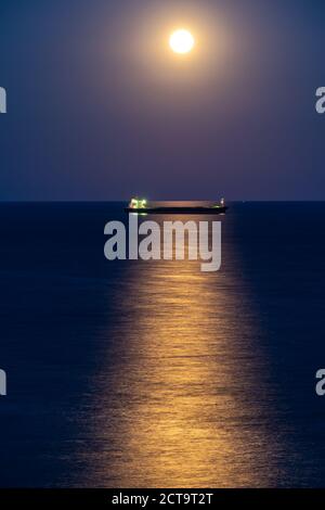 Frachtschiff bei Nacht mit Vollmond über dem Horizont Stockfoto